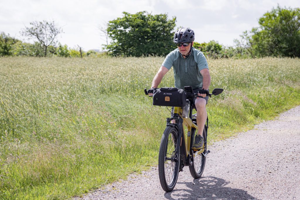 Älterer Mann mit Helm radelt mit Fronttasche auf sonnigem Weg.