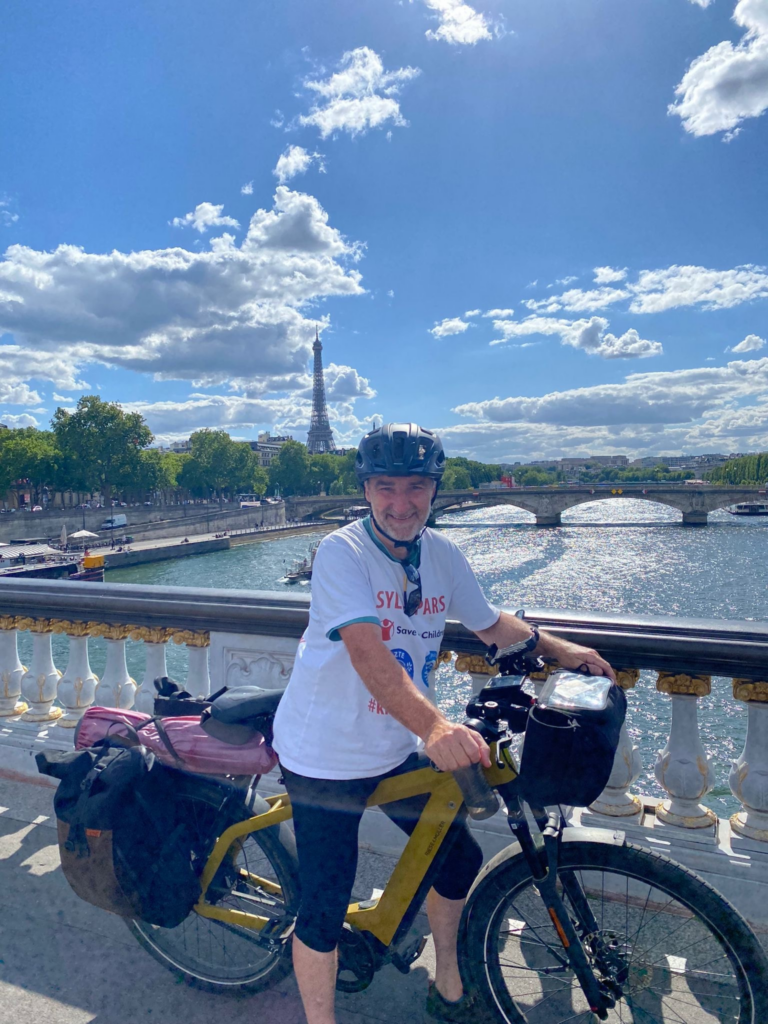 Behelmter Radfahrer mit beladenem Fahrrad auf einer Pariser Brücke, dahinter der Eiffelturm.
