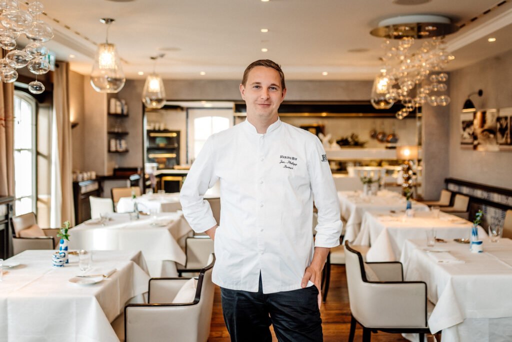 Chef in a white jacket in the bright dining room.