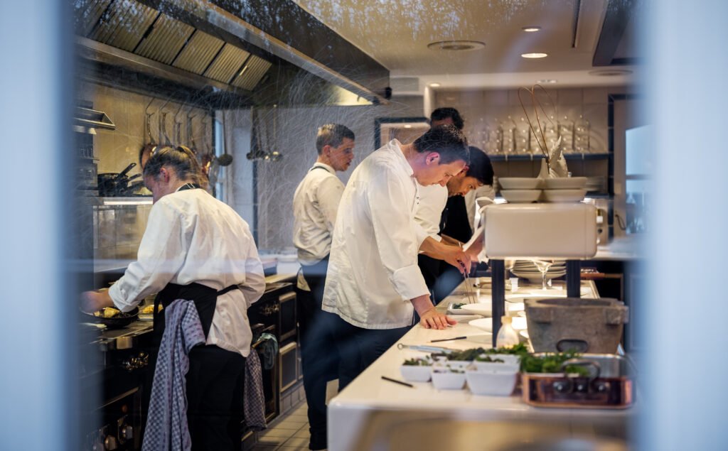 Die offene Küche vom Söl'ring Hof aus der seitlichen Perspektive während viele Köche anrichten und der Service läuft. Küchenchef Jan-Philipp Berner steht ebenfalls am Pass.