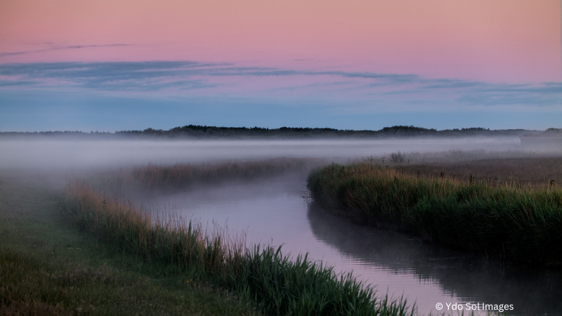 Ein ruhiger Fluss windet sich bei Sonnenuntergang durch nebliges Gras.