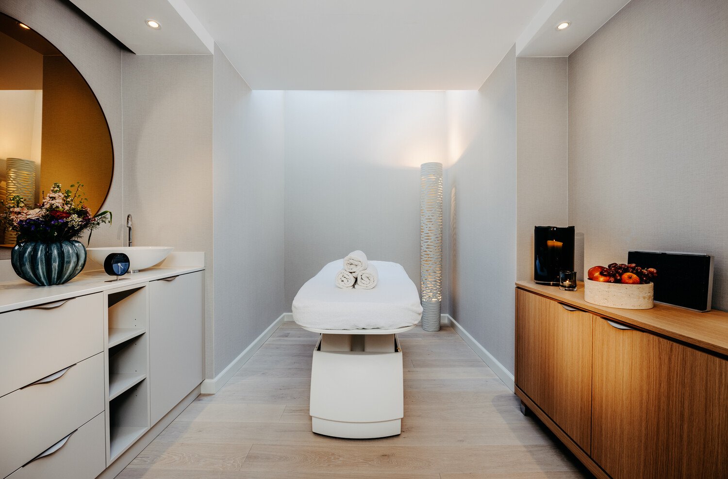 Eine weiße Massageliege steht in der Mitte des Massage Raumes mit kleinen Regalen auf beiden Seiten an den Wänden
