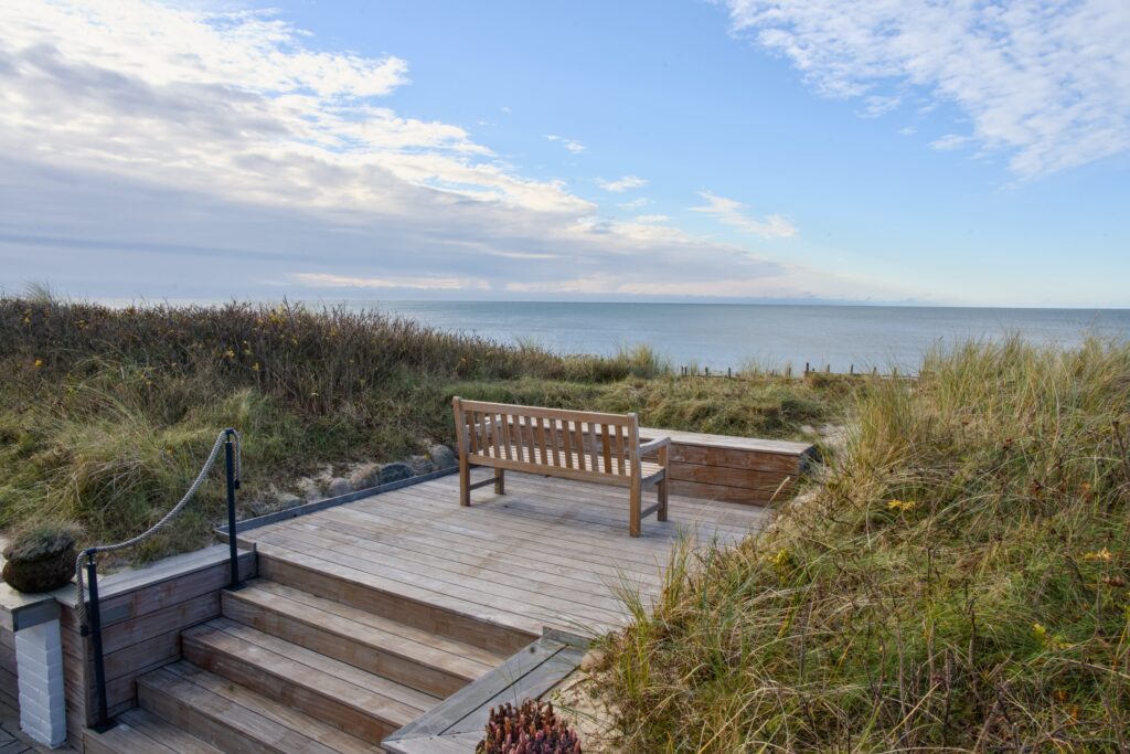 Bench on deck overlooks sand dunes, ocean; partly cloudy sky.