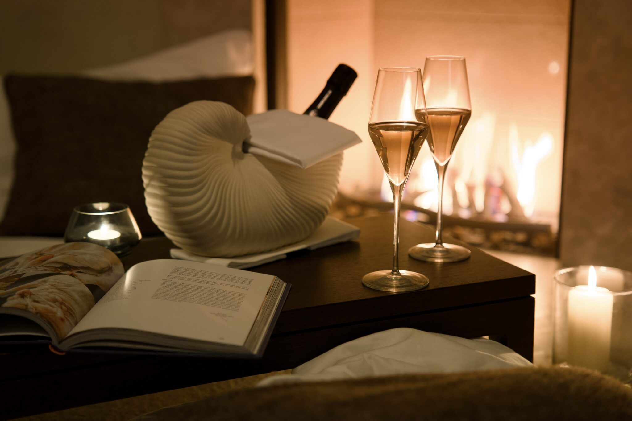 Champagne, wine, book, and candles on table before fireplace—cozy.