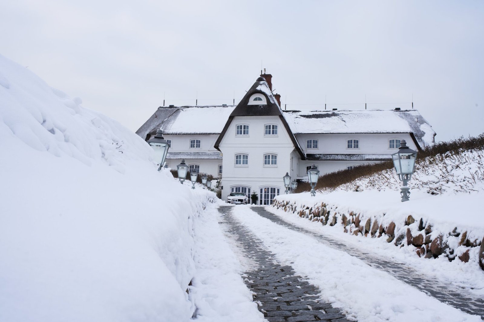 Weißes Haus, steiles Dach, verschneiter Weg, Straßenlampen, bewölkter Himmel.
