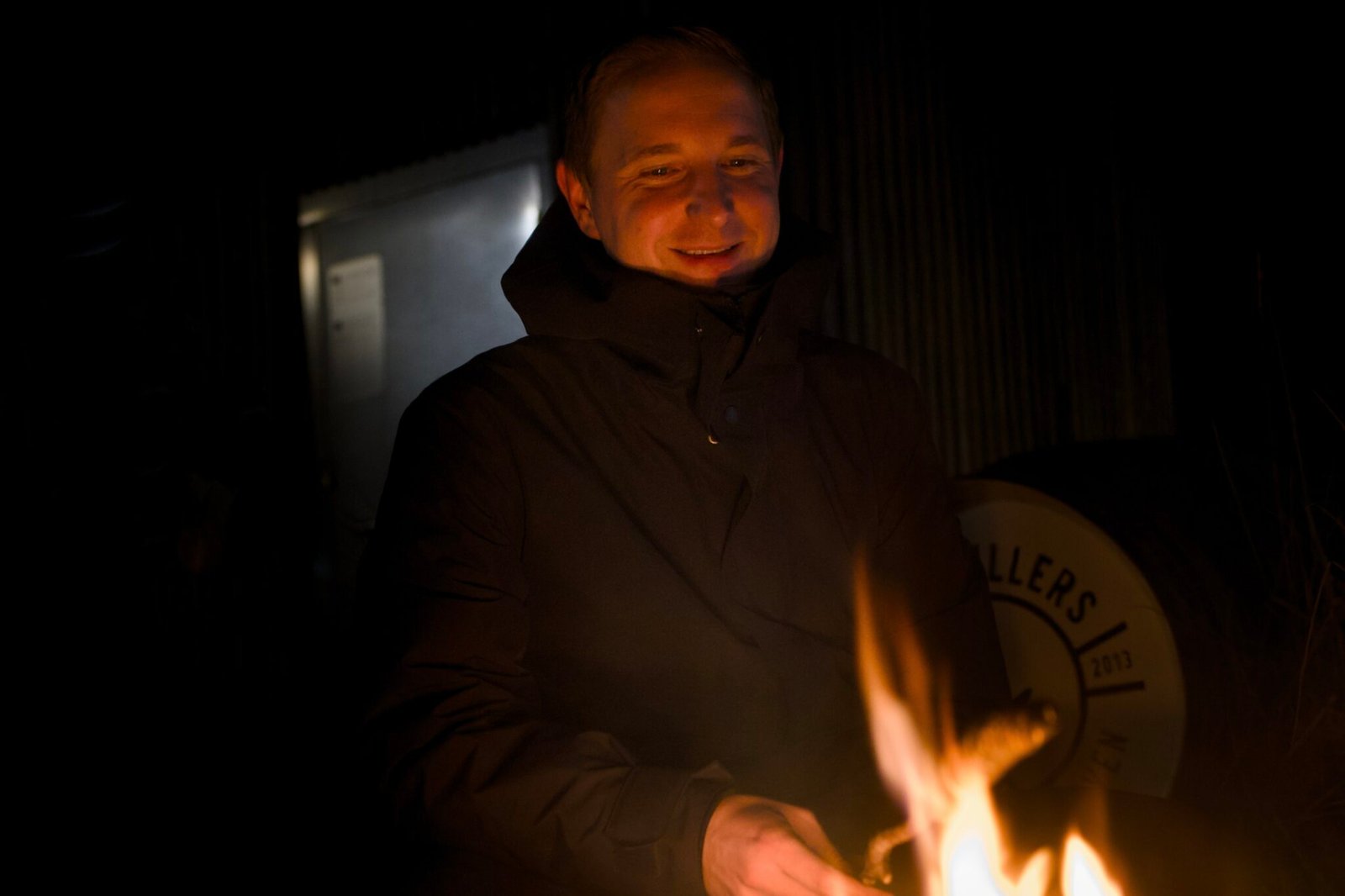 Person in dunkler Jacke am Lagerfeuer, dahinter Metallwand und Fass.