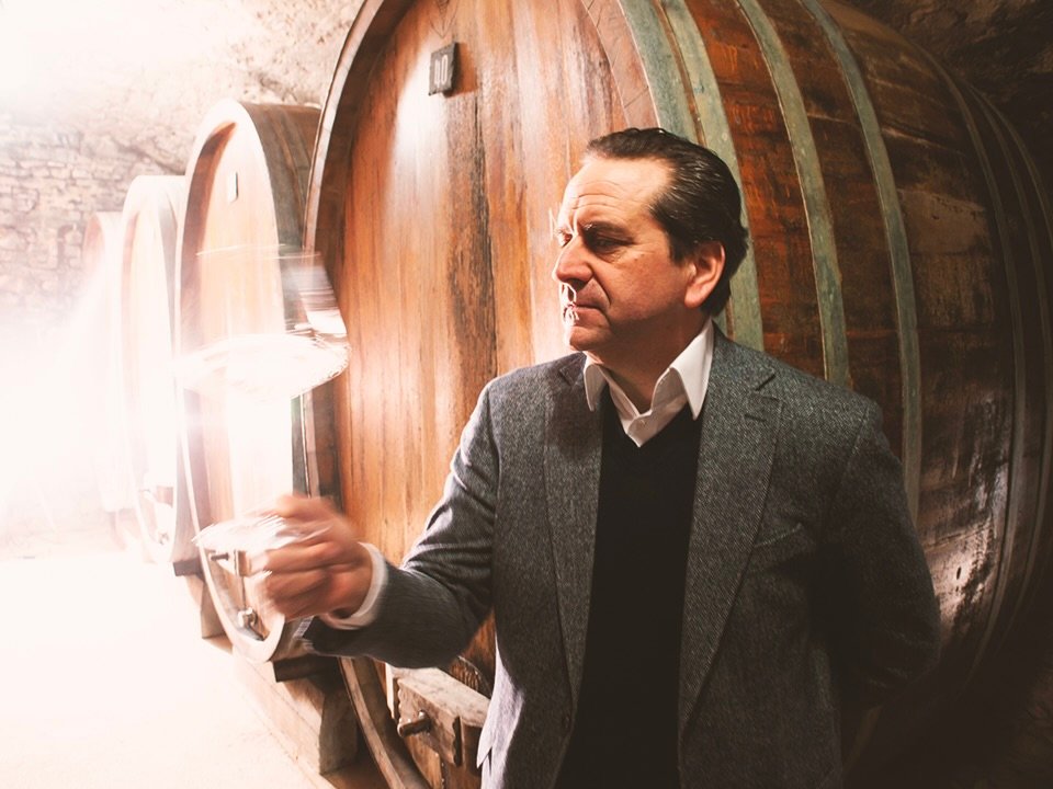 Man in suit examines white wine before cellar barrels, warm lighting.