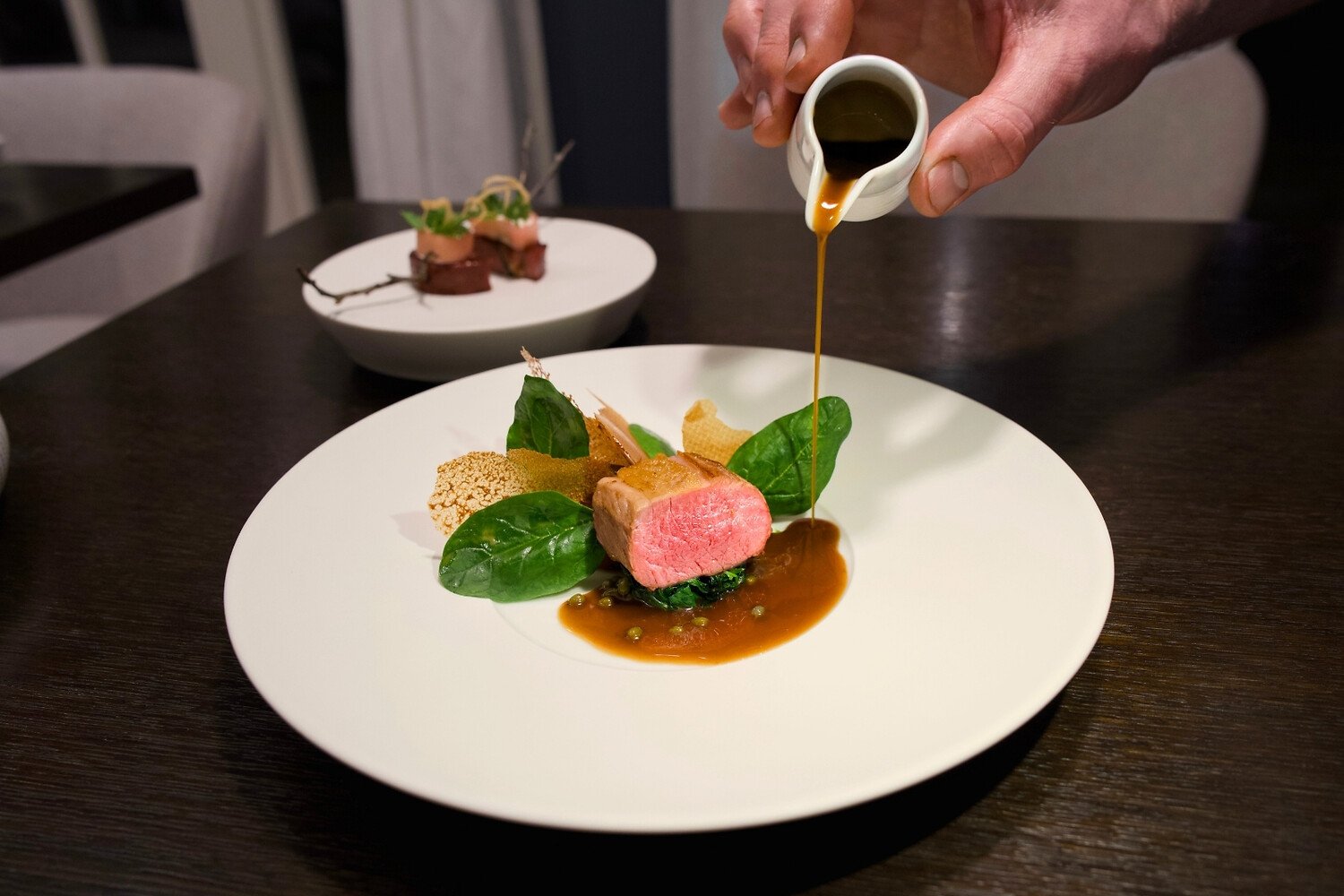 Hand pours sauce over seared meat and greens on plate.