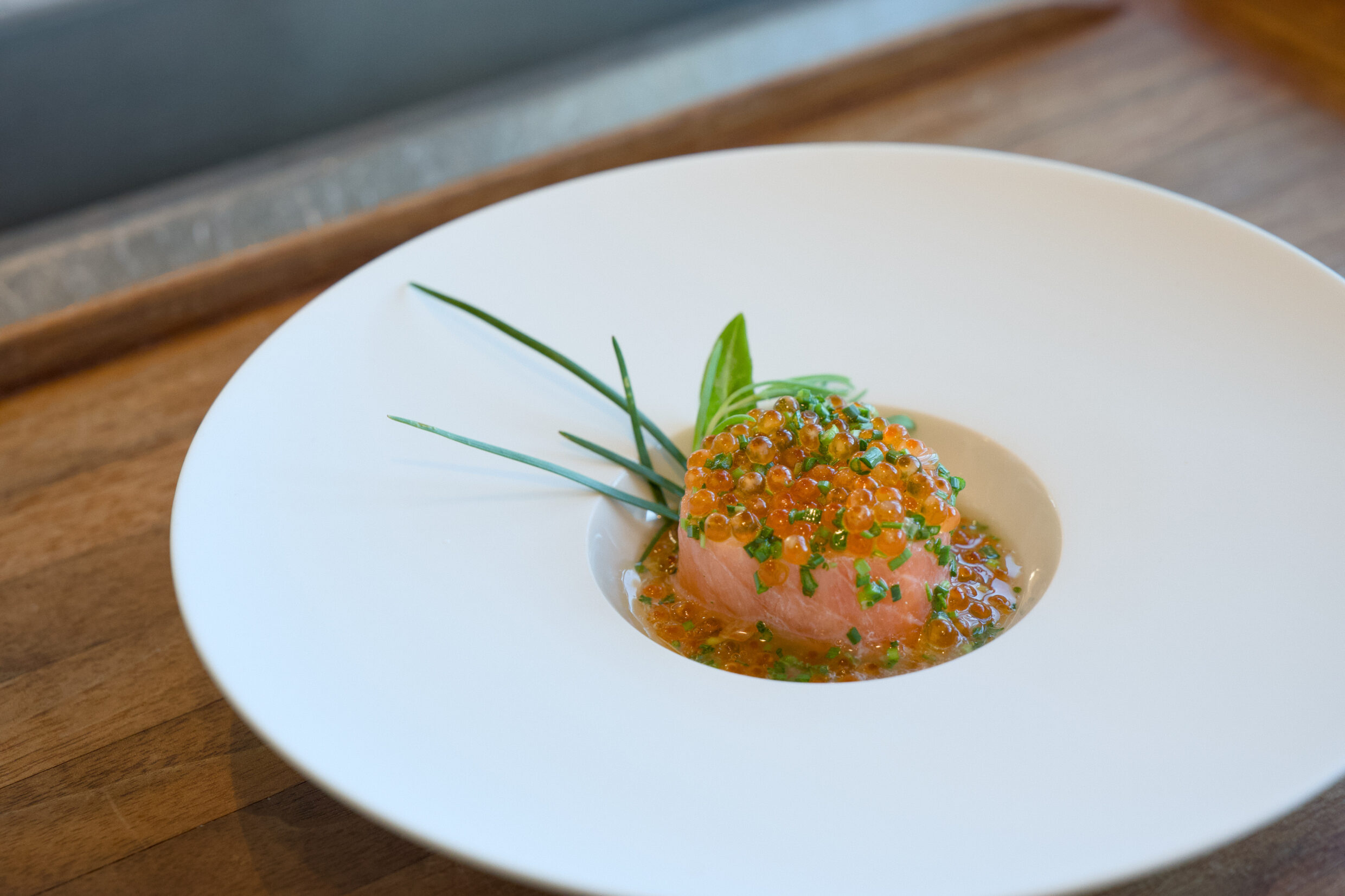Roher Fisch mit Orangenrogen und Kräutern auf einem runden Teller.