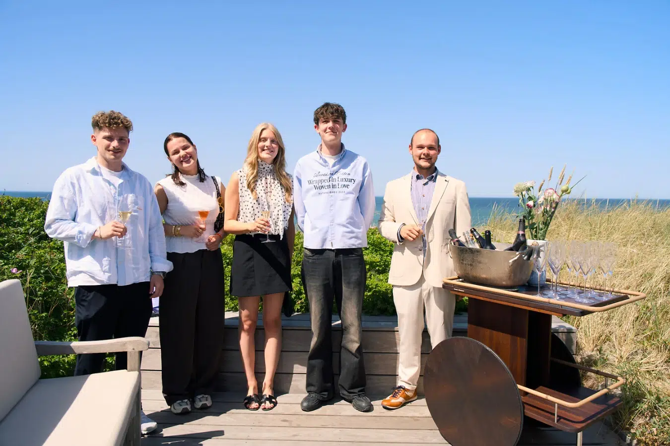 Fünf Personen in halbformeller Kleidung stehen auf einer Terrasse am Strand.