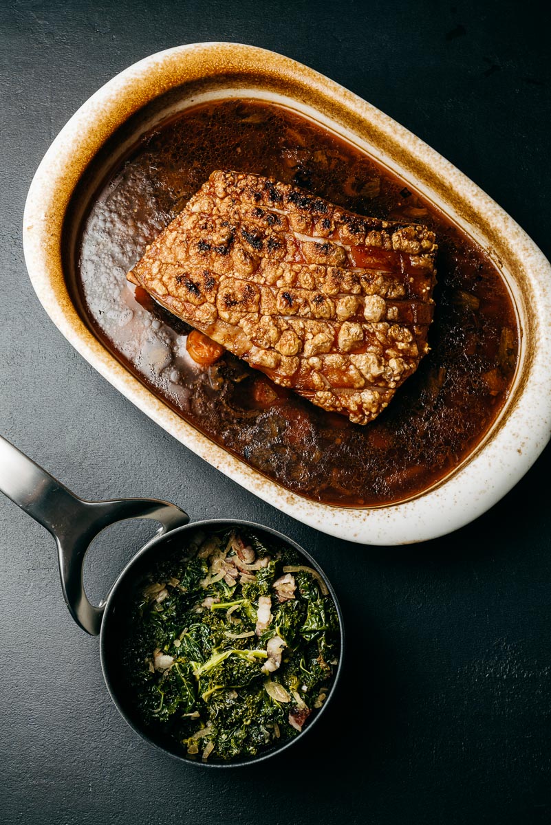 Roast pork belly with crackling, greens, and bacon on dark surface.