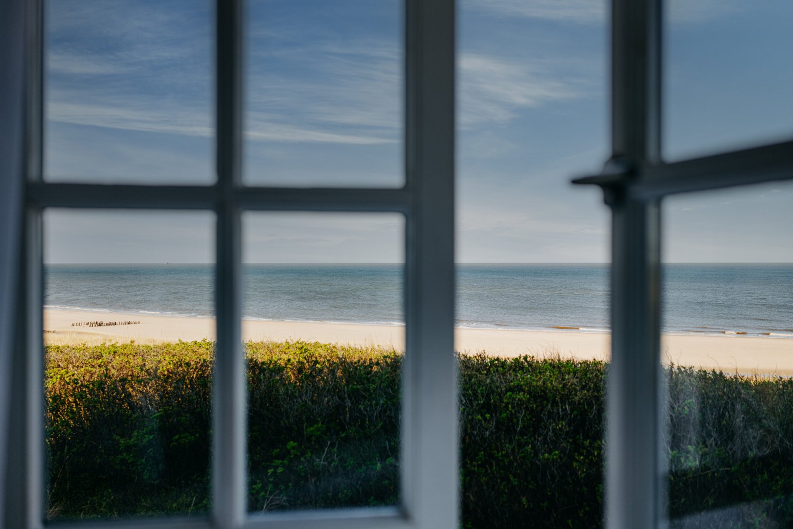 Sandstrand, Sträucher und das Meer, gesehen durch ein Fenster mit weißem Rahmen.