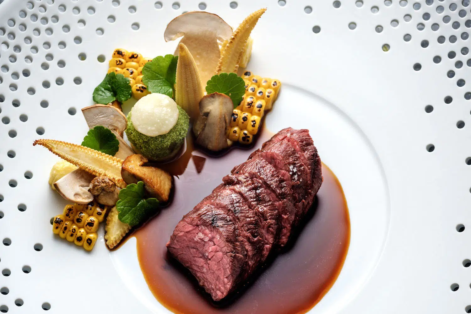 Rare beef with corn, mushrooms, greens, sauces on patterned plate.