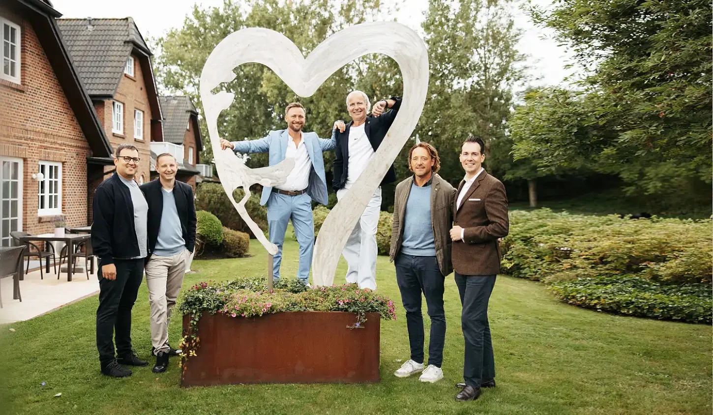 Sechs Männer posieren in einem Garten vor einer weißen Herzskulptur.